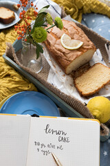 Lemon cake recipe written in a notebook. A delicious cake with a laid slice is at the background.