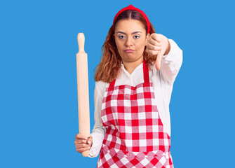 Young latin woman wearing apron holding kneader with angry face, negative sign showing dislike with...