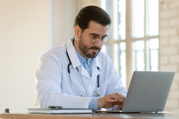Fototapeta premium Advancing in skills. Inspired concentrated millennial doctor intern qualified specialist using computer to read medical literature online, asking questions on professional forums to specify diagnosis