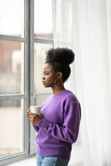 Pensive African American millennial biracial woman with trendy jewelry earrings wear purple sweater thinking, looking through the window, holding white mug, drinking hot coffee or tea. 