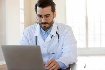 Fototapeta na wymiar Thoughtful focused male physician working on laptop in hospital office counseling online, providing virtual appointment, studying new academic research by his speciality, thinking on electronic report
