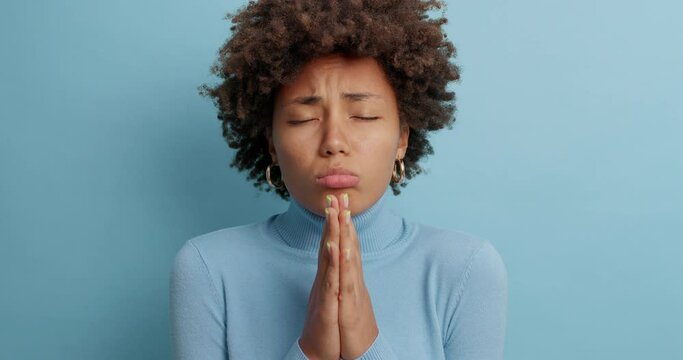 Sad dark skinned woman looks with pleading expression asks to forgive her for making mistake apologizes and purses lips dressed in casual turtleneck isolated over blue background. Forgiveness