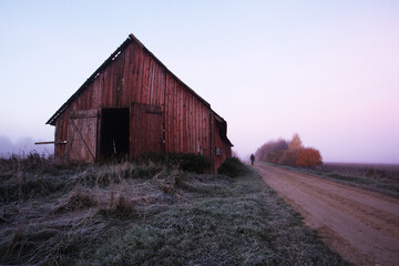 An old red wooden barn next to a small dirt road on a misty autumn morning in Estonian countryside. 