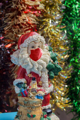 A figure of Santa Claus standing with a bag full of toys wears a mask that covers his nose and mouth against a background of colorful tinsel. Coronavirus, Christmas, and gifts.