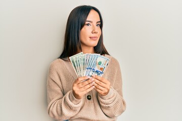 Young beautiful hispanic girl holding dollars smiling looking to the side and staring away thinking.