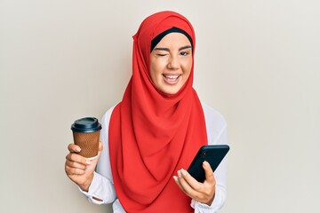 Young beautiful hispanic girl wearing islamic hijab drinking a coffee usign smartphone winking looking at the camera with sexy expression, cheerful and happy face.