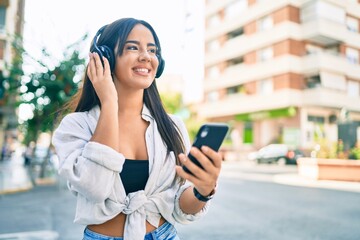 Young hispanic girl smiling happy using smartphone and headphones at the city.