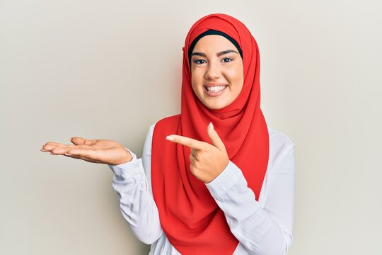 Young beautiful hispanic girl wearing traditional islamic hijab scarf amazed and smiling to the camera while presenting with hand and pointing with finger.