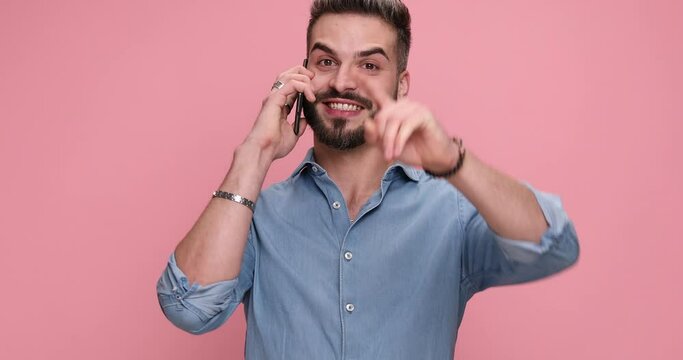 casual young man talking on the phone, smiling, pointing finger on pink background