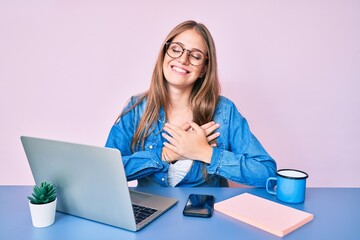 Young blonde girl working at the office drinking a cup of coffee smiling with hands on chest with closed eyes and grateful gesture on face. health concept.