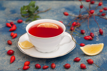 Cup of rosehip herbal tea with rose hip fruits and lemon on blue rustic background. Dogrose autumn or winter healthy tea.