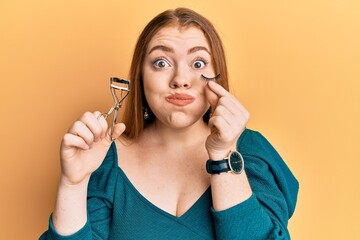 Young beautiful redhead woman holding eyelash curler and fake lashes puffing cheeks with funny face. mouth inflated with air, catching air.