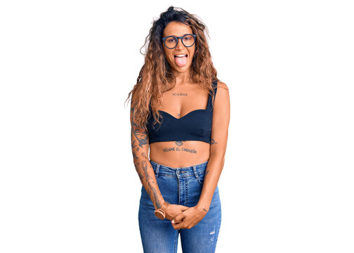 Young hispanic woman with tattoo wearing casual clothes and glasses sticking tongue out happy with funny expression. emotion concept.