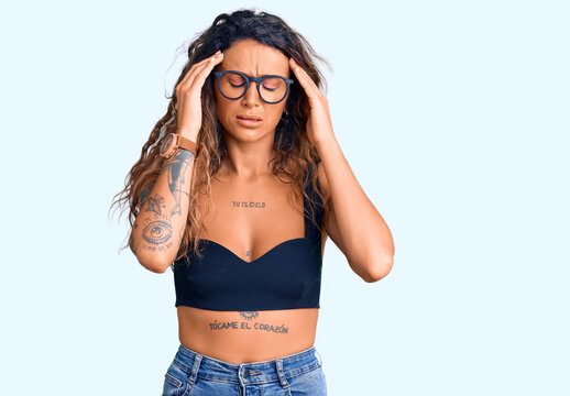 Young hispanic woman with tattoo wearing casual clothes and glasses with hand on head for pain in head because stress. suffering migraine.