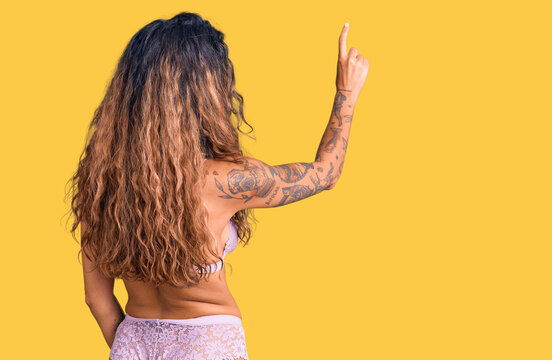 Young hispanic woman with tattoo wearing lingerie posing backwards pointing ahead with finger hand