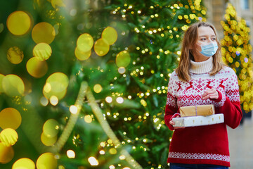 Happy young girl wearing face mask in holiday sweater with pile of holiday gifts