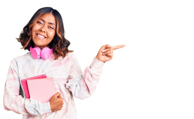 Young beautiful mixed race woman holding books wearing headphones smiling happy pointing with hand and finger to the side