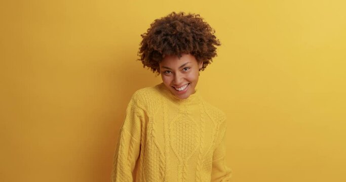 Optimistic joyous young woman with Afro hair dances carefree and raises arms dressed in casual clothing celebrates something spends free time on disco party isolated over yellow studio background