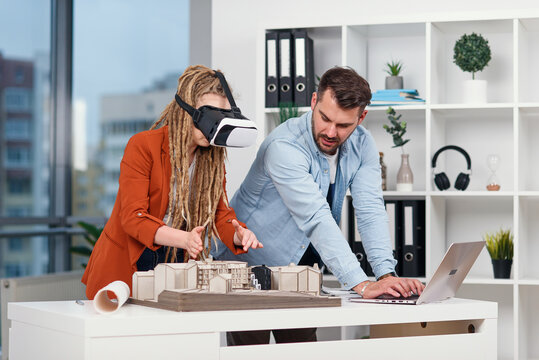 Skillful male architect working on laptop while his female colleague in VR glasses analysing mock-up of future building.