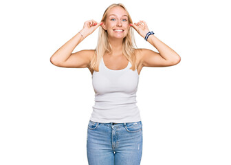 Young blonde girl wearing casual style with sleeveless shirt smiling pulling ears with fingers,...