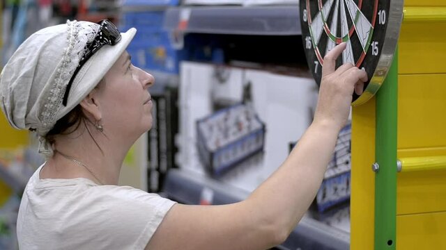 Lady chooses a dart board in the store. A customer inspects a Board for playing butt. The lady is wearing a hat and sunglasses.