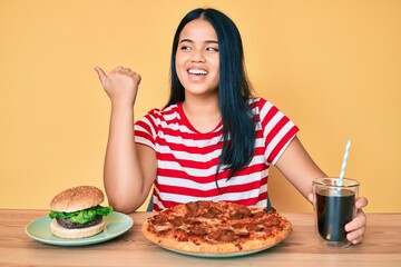 Young beautiful asian girl eating fast food pizza and burger pointing thumb up to the side smiling happy with open mouth