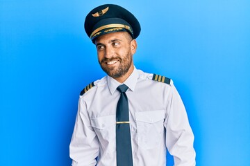 Handsome man with beard wearing airplane pilot uniform looking away to side with smile on face, natural expression. laughing confident.