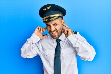 Handsome man with beard wearing airplane pilot uniform covering ears with fingers with annoyed expression for the noise of loud music. deaf concept. © Krakenimages.com