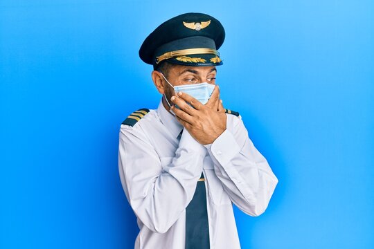 Handsome Man With Beard Wearing Airplane Pilot Uniform Wearing Safety Mask Shocked Covering Mouth With Hands For Mistake. Secret Concept.