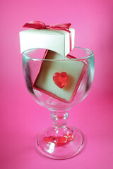 Romantic holiday greeting card. Beautiful glass with a red heart shape and a gift on a pink background. Valentine's day, wedding
