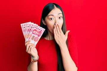 Young chinese woman holding 10 yuan chinese banknotes covering mouth with hand, shocked and afraid...
