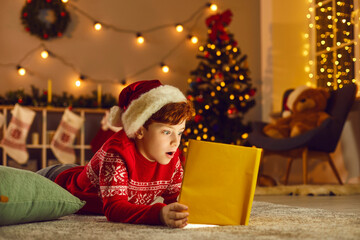 Little boy opens magic book and reads wonderful stories and fairy tales on Christmas Eve at home