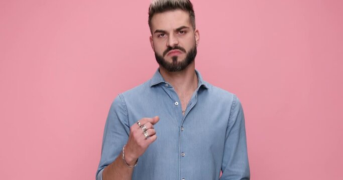 attractive casual man feeling desperate over someone, being aggresive, pointing his finger and threatening, showing his fist, running his hands trough his head against pink background