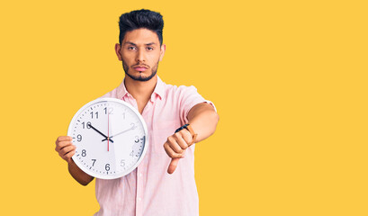 Handsome latin american young man holding big clock with angry face, negative sign showing dislike...