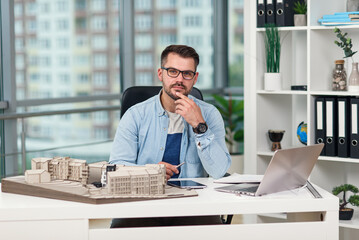 Concentrated successful bearded architect at eyeglasses sits at his workplace with a layout of future buildings.
