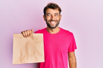 Handsome man with beard holding take away paper bag looking positive and happy standing and smiling with a confident smile showing teeth