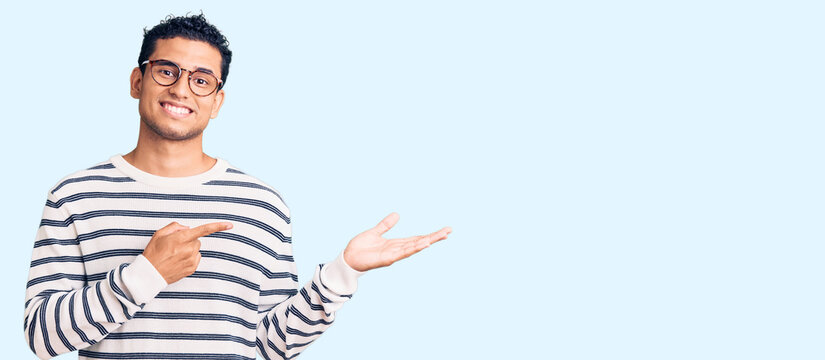 Hispanic handsome young man wearing casual clothes and glasses amazed and smiling to the camera while presenting with hand and pointing with finger.