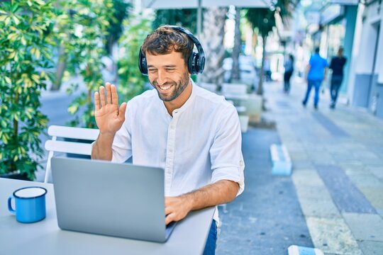 Handsome man with beard wearing casual white shirt on a sunny day working using laptop and wearing headphones and waving to videocall at cafeteria