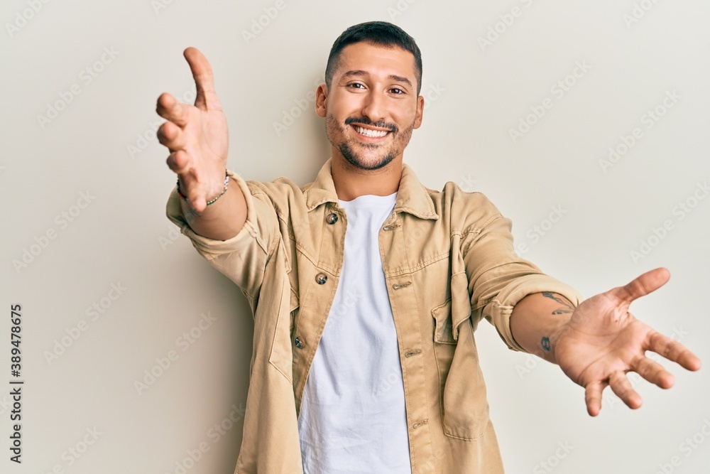 Wall mural Handsome man with tattoos wearing casual brown denim jacket looking at the camera smiling with open arms for hug. cheerful expression embracing happiness.