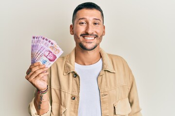 Handsome man with tattoos holding 50 mexican pesos banknotes looking positive and happy standing and smiling with a confident smile showing teeth