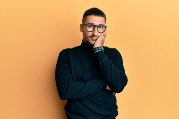 Handsome man with tattoos wearing turtleneck sweater and glasses thinking looking tired and bored with depression problems with crossed arms.