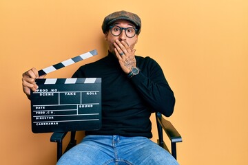 Handsome man with tattoos holding video film clapboard sitting on director chair covering mouth...