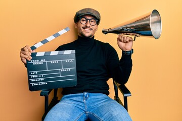 Handsome man with tattoos holding video film clapboard and louder smiling with a happy and cool smile on face. showing teeth. - obrazy, fototapety, plakaty
