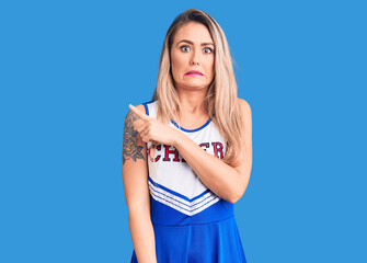 Young beautiful blonde woman wearing cheerleader uniform pointing aside worried and nervous with...
