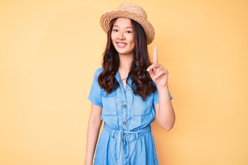 Young beautiful chinese girl wearing summer hat showing and pointing up with finger number one while smiling confident and happy.