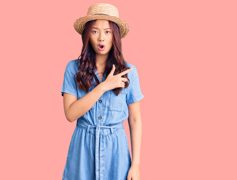 Young beautiful chinese girl wearing summer hat surprised pointing with finger to the side, open mouth amazed expression.