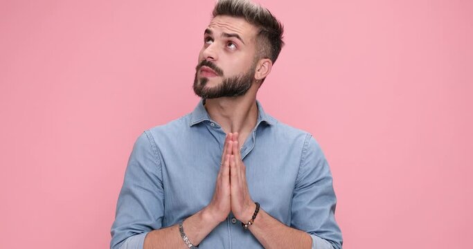 serious casual  man praying with his hands together for better days and feeling troubled against pink background