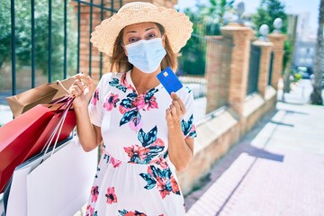 Middle age hispanic woman wearing medical mask holding shopping bags and credit card at city