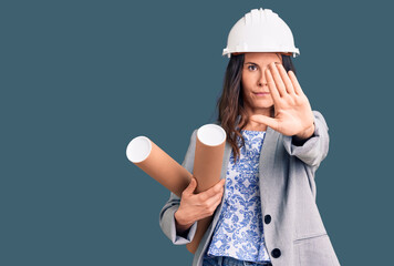 Young beautiful brunette woman wearing architect hardhat holding blueprint with open hand doing stop sign with serious and confident expression, defense gesture