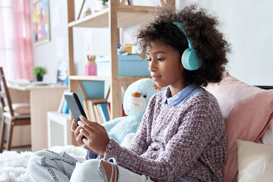 African american child kid girl wearing headphones holding phone sitting on bed. Cute black kid using smartphone tech listening course watching video class elearning on cellphone in bedroom at home.